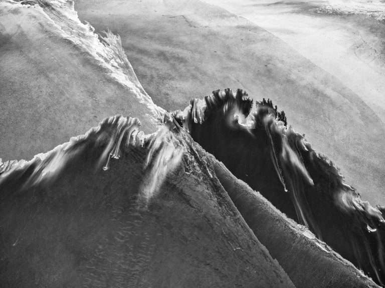 Black-and-white aerial photograph of Kati Thanda–Lake Eyre showing dark water channels cutting through pale salt flats, forming wave-like ridges and flowing textures that resemble sculpted rock or liquid ink.