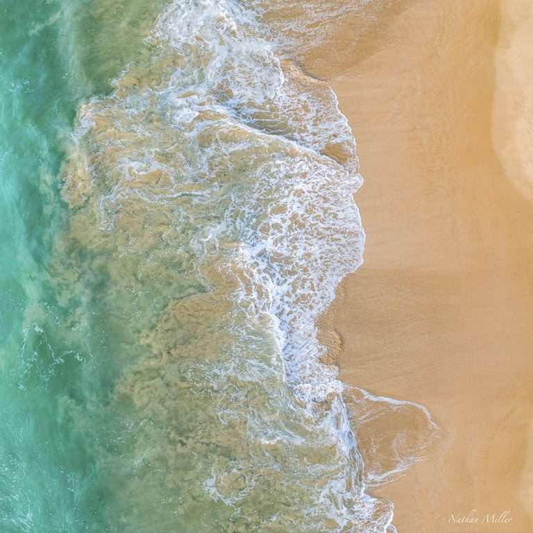 Aerial view of turquoise ocean water washing onto warm golden sand, with white foam forming soft, flowing patterns along the shoreline.